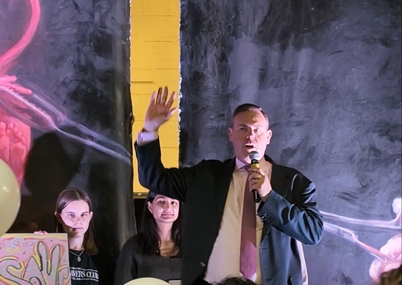 Mike wears a suit jacket and speaks into a microphone, waving one hand in the air.