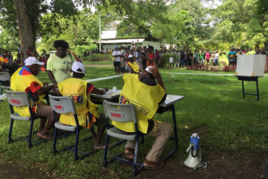 Officials in yellow vests sit at a table as voters line up.