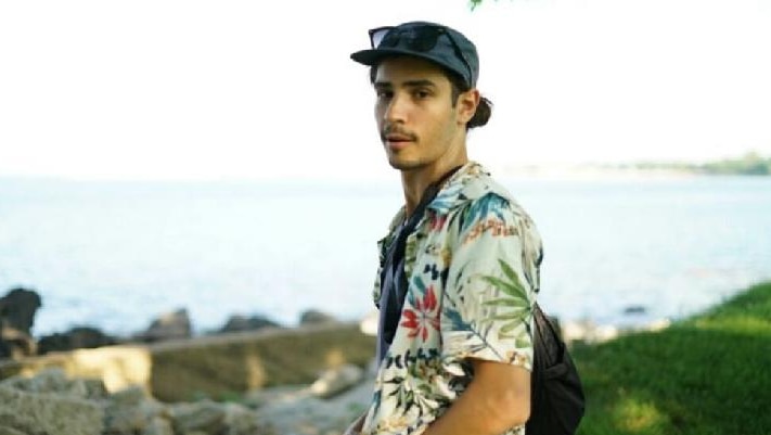 A photo of a young man wearing a hat with his sunglasses on it, and a floral shirt.