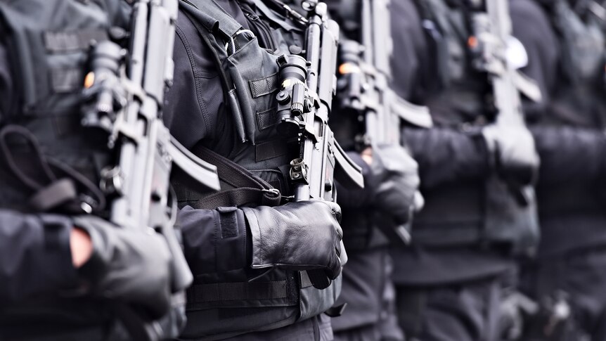 A line of soldiers wearing black gloves and holding automatic machine guns.