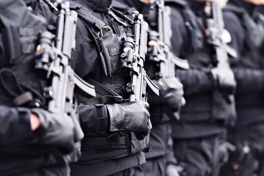 A line of soldiers wearing black gloves and holding automatic machine guns.