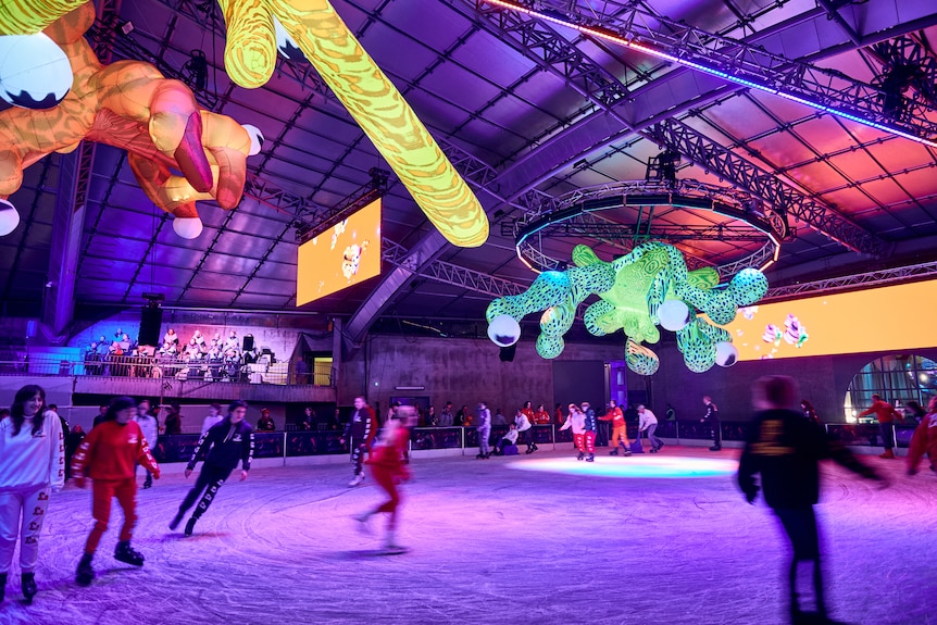 Brightly coloured inflatable sculptures hang from the roof of an ice-skating rink, as a choir performs in the bleachers