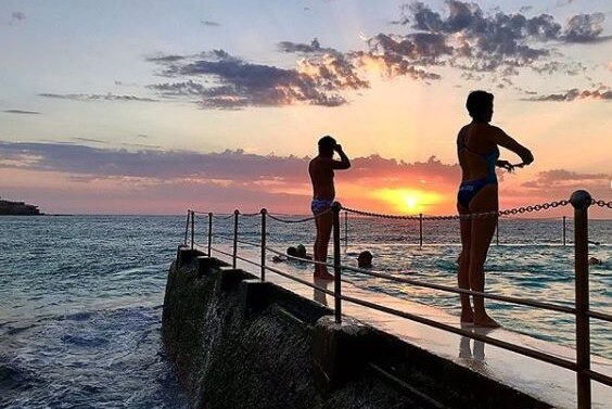 Two people in swimming costumes stand at the edge of an ocean pool