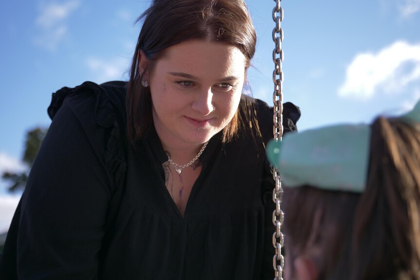 A dark-haired woman looks at her young daughter, who's facing her in a swing