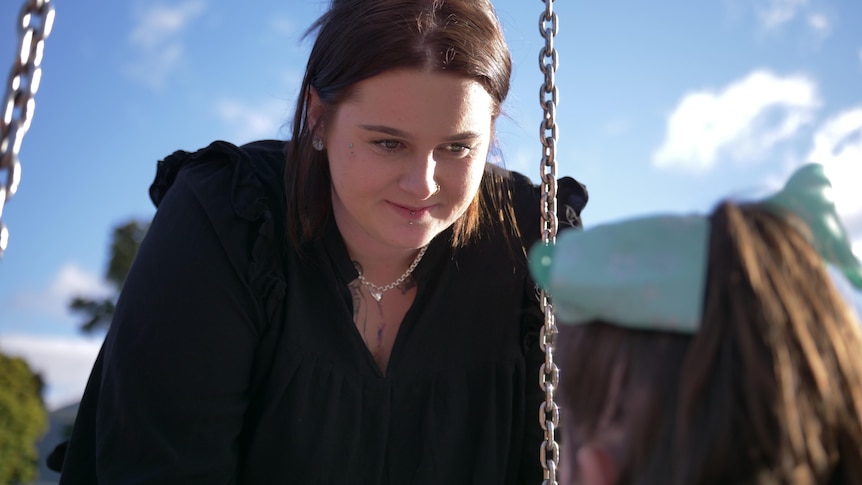 A dark-haired woman looks at her young daughter, who's facing her in a swing