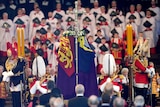 Queen Elizabeth's coffin is draped with a flag and guarded by several guards, in front of a choir