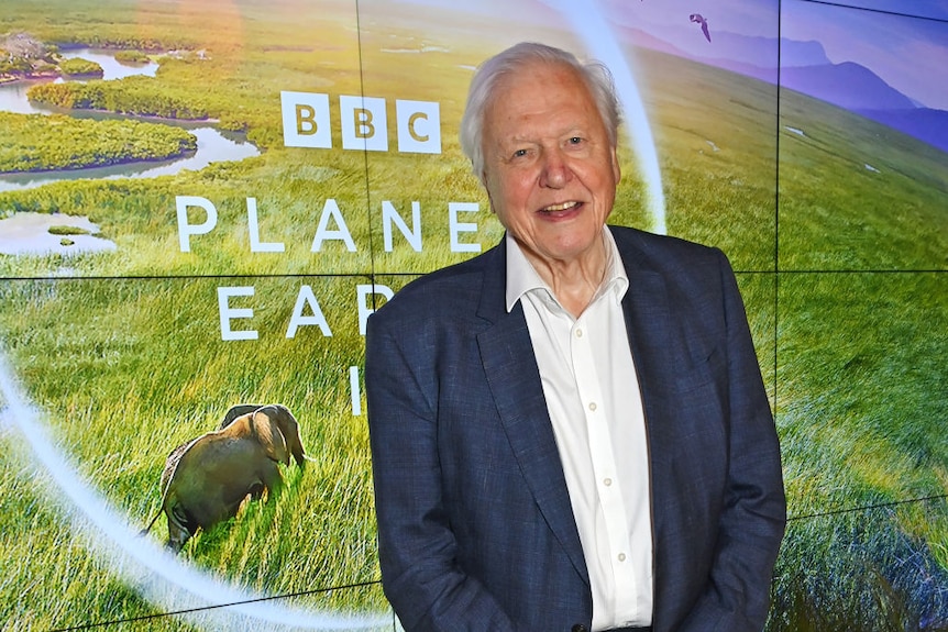 A smiling older man in white shirt and dark blue suit standing in front of a screen with the text Planet Earth III