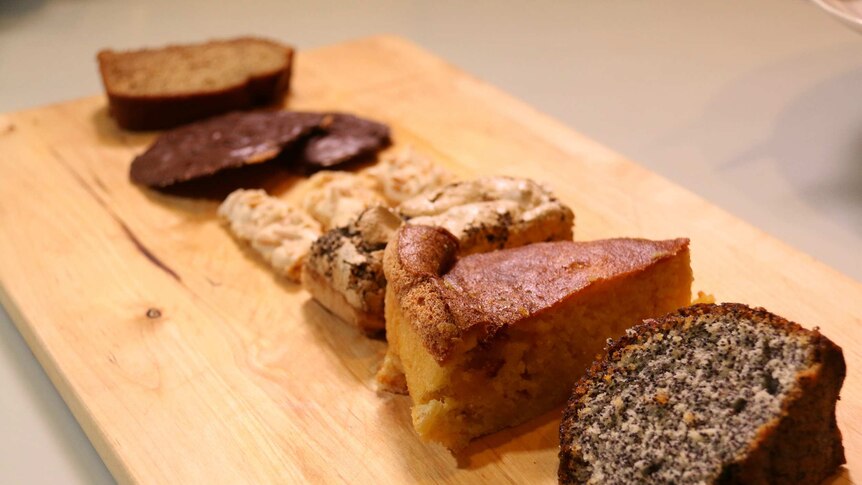 Slices of six different types of cake are on a wooden chopping board.