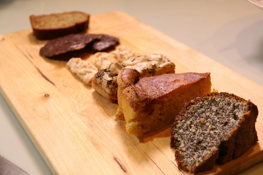 Slices of six different types of cake are on a wooden chopping board.