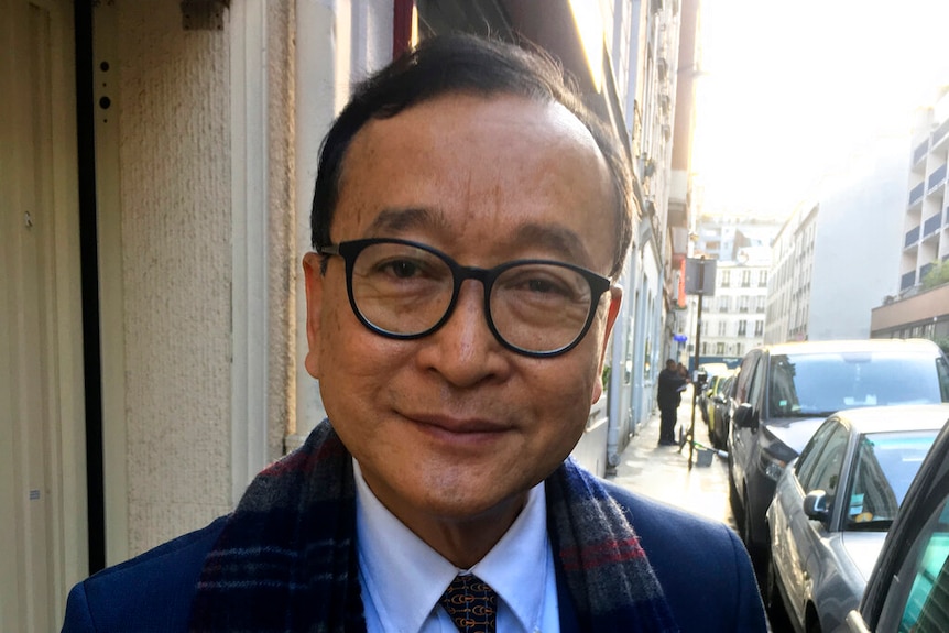 A close portrait of Sam Rainsy wearing a navy suit and chequered scarf on a narrow Parisian street.