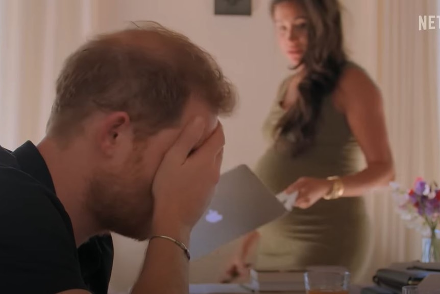 Prince Harry puts his head in his hands as Meghan stands behind him