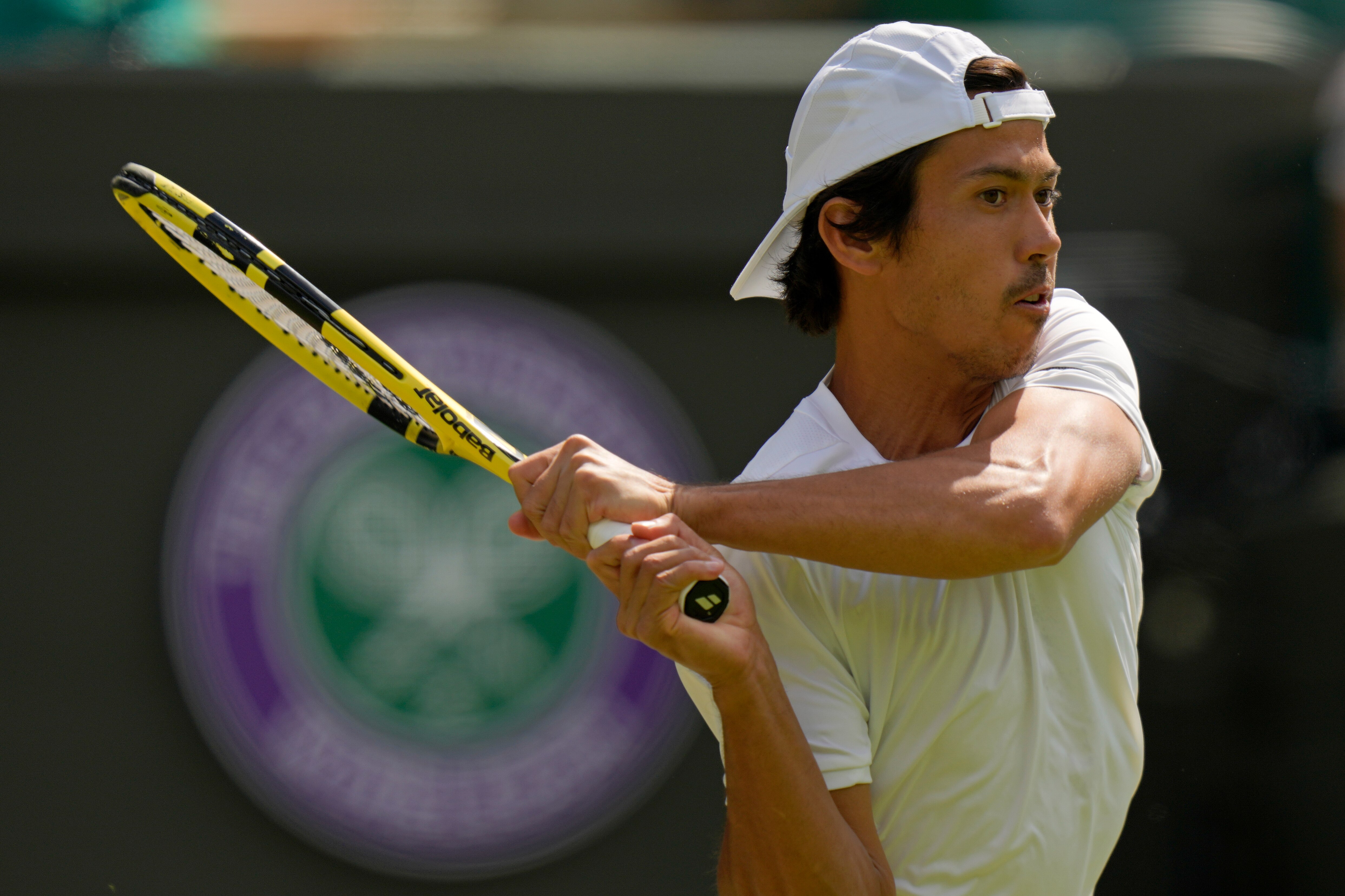 Australian tennis player Jason Kubler, wearing a white shirt and backwards cap, plays a backhand in front of Wimbledon's logo.