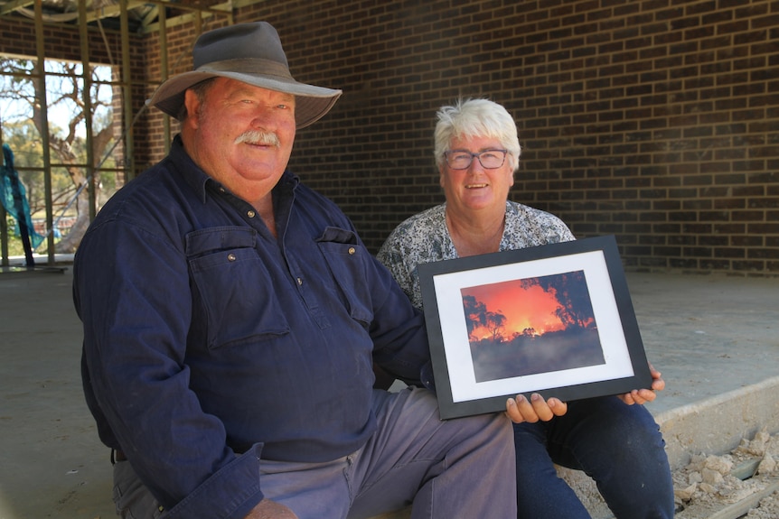 A man and a woman sit in a garage that's being built holding an image of a bushfire on their property. 
