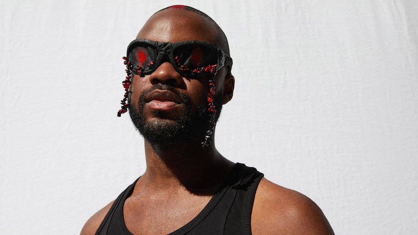 Genesis Owusu stands in front of a white background with black and red sunglasses on and a black singlet