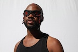 Genesis Owusu stands in front of a white background with black and red sunglasses on and a black singlet