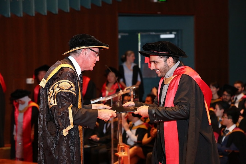 A man in a college gown and hat gets his PhD from another man in a college gown and hat.