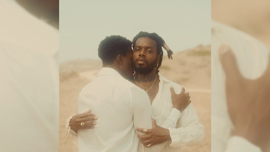 serpentwithfeet embraces a man in the desert. they both wear white. the word DEACON is tattooed across his neck