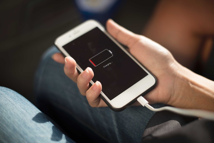 A hand holds a white mobile phone that is plugged in and showing a charging icon on its screen.