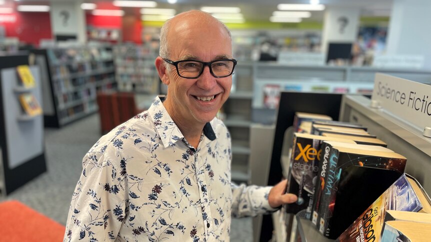 A middle aged man wearing glasses and a patterned shirt stands among shelves of books in a library.