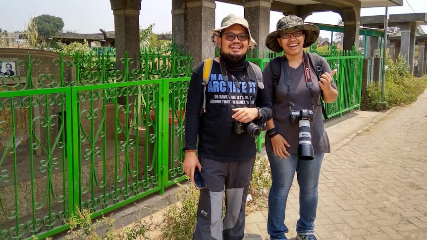 A man and woman stand outside a cemetery. They are both wearing broad-brimmed hats and carrying cameras.