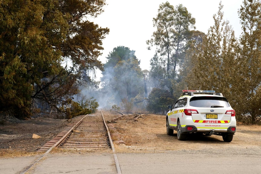 A fire car parked by a railway line where fallen trees smoke with embers.