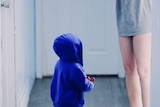 Child in blue hooded jumper stands in hallway next to mum in long grey t-shirt to depict childhood stonewalling and its effects.