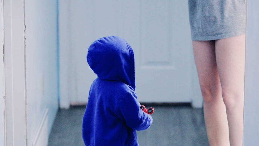 Child in blue hooded jumper stands in hallway next to mum in long grey t-shirt to depict childhood stonewalling and its effects.