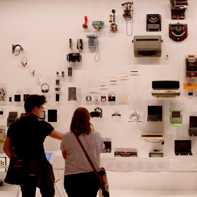 A series of writing, timekeeping, and computing are against a white gallery wall, with three people looking on behind labels.