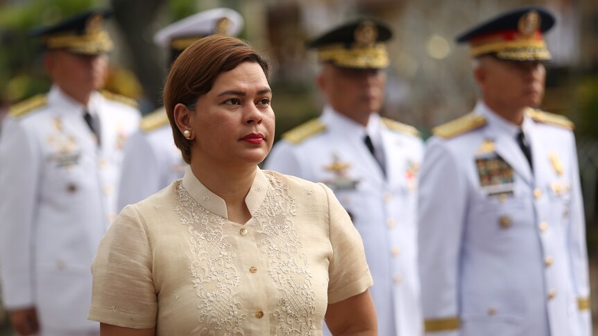 Sara Duterte stands in front of military officers in white as she gazes into the distance while wearing ceremonial dress.