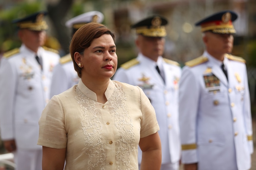 Sara Duterte stands in front of military officers in white as she gazes into the distance while wearing ceremonial dress.