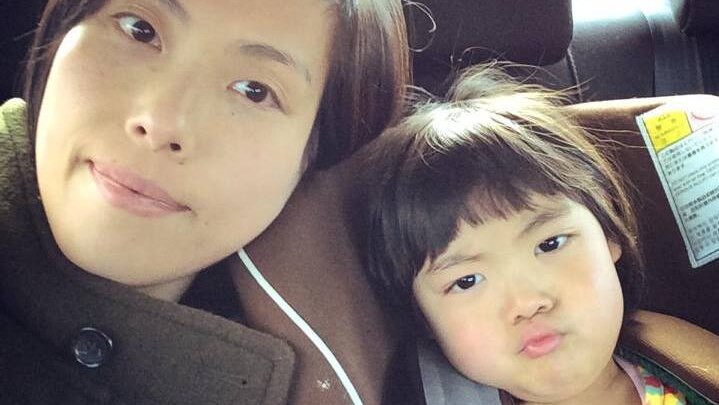 Kaoru Okano smiles at the camera, with her daughter in a car seat next to her.