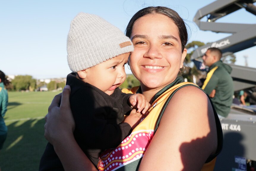Young footy player holding a little baby boy in her guernsey.