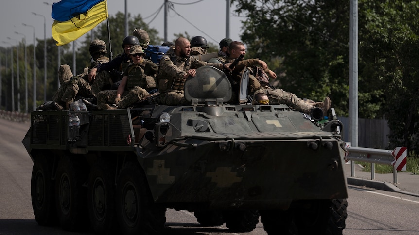 a dozen ukrainian soldiers sit on a troop with a ukrainian flag, as it rolls down a main street