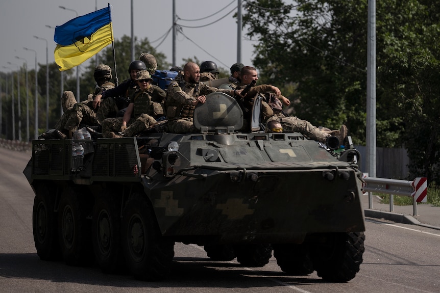 a dozen ukrainian soldiers sit on a troop with a ukrainian flag, as it rolls down a main street