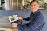 Man sits at table, looking at iPad with a house advertisement on the screen.