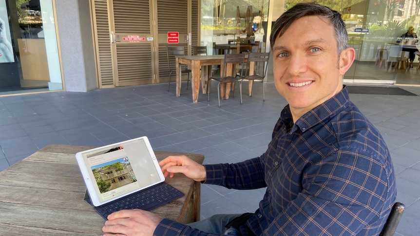 Man sits at table, looking at iPad with a house advertisement on the screen.