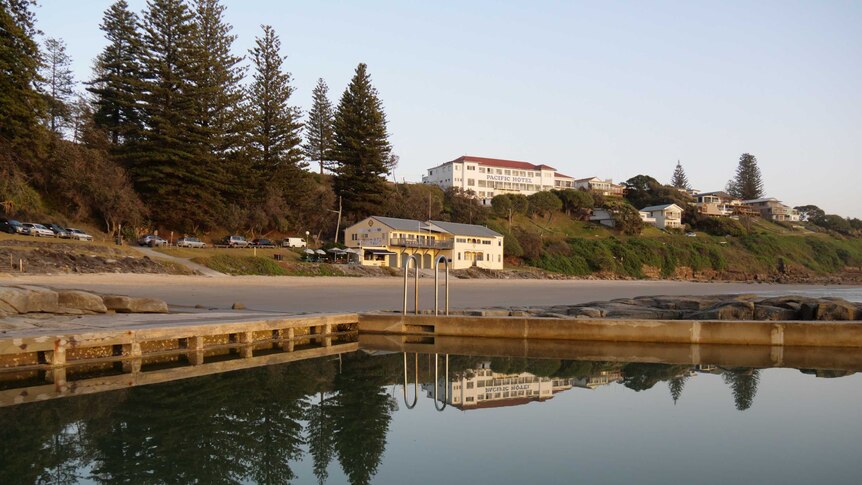 An ocean pool glistens at dawn with a glacially flat surface, in the background is a clifftop pub signed 'Pacific Hotel'.