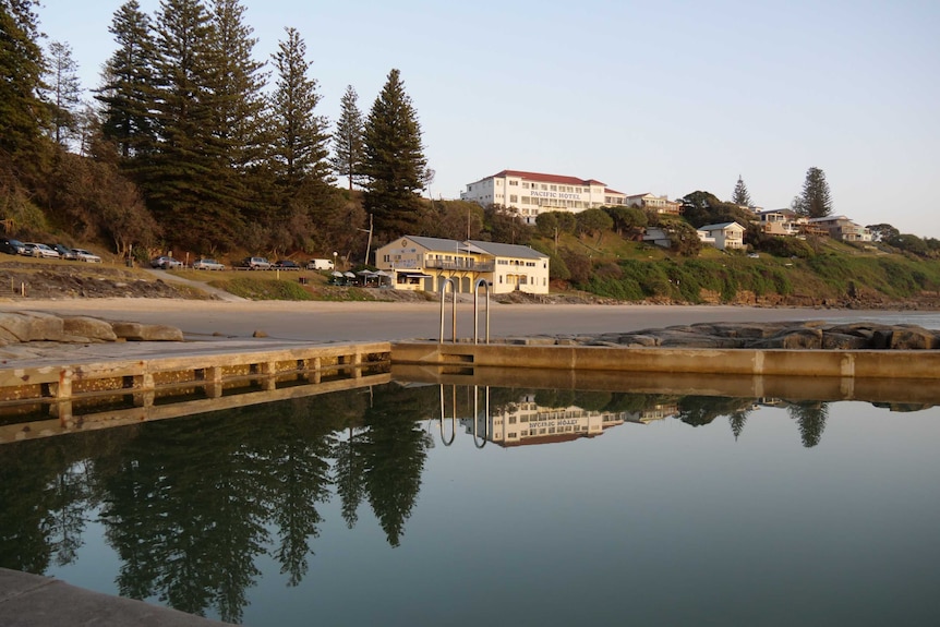 An ocean pool glistens at dawn with a glacially flat surface, in the background is a clifftop pub signed 'Pacific Hotel'.