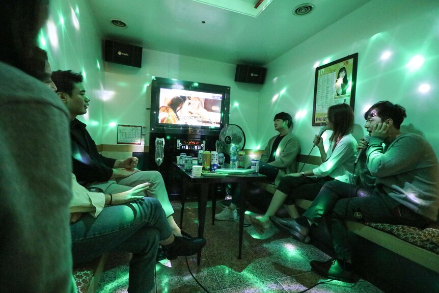 students gather around a television in a green lit karaoke room