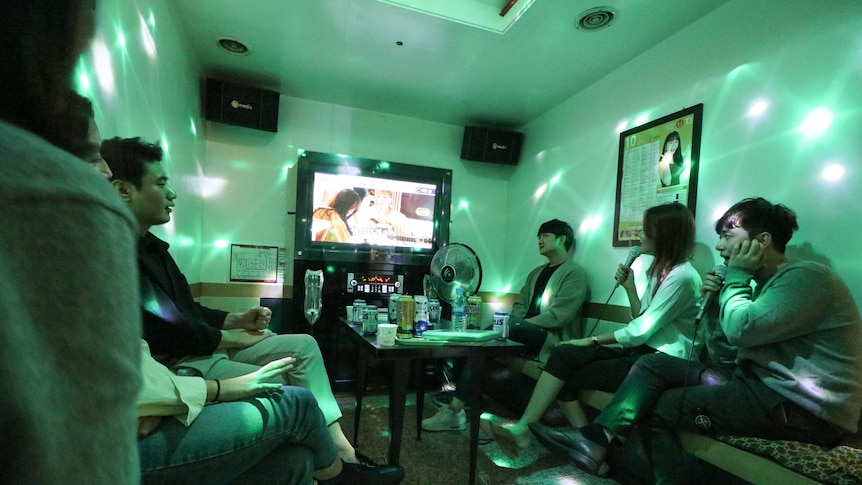 students gather around a television in a green lit karaoke room