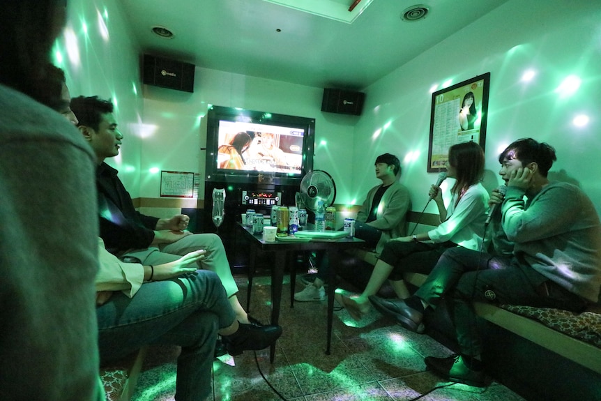 students gather around a television in a green lit karaoke room