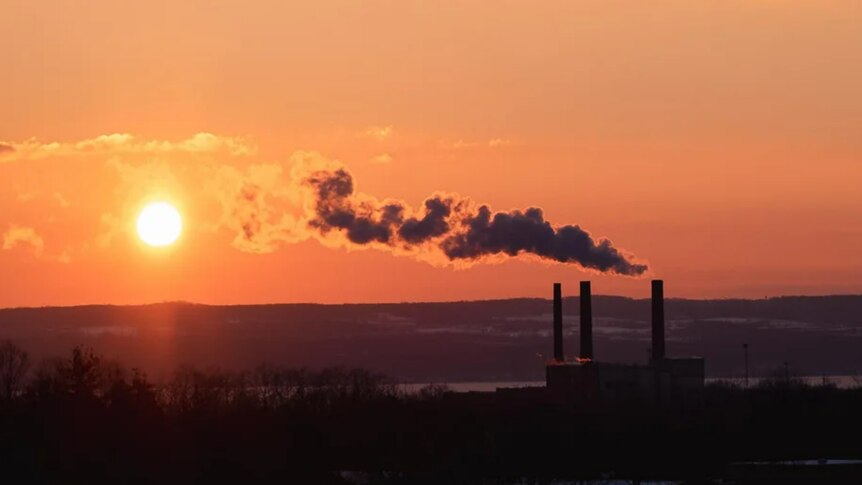 A factory with three smoke stacks pumps smoke into an orange sky at dawn set against a lake and rolling hills