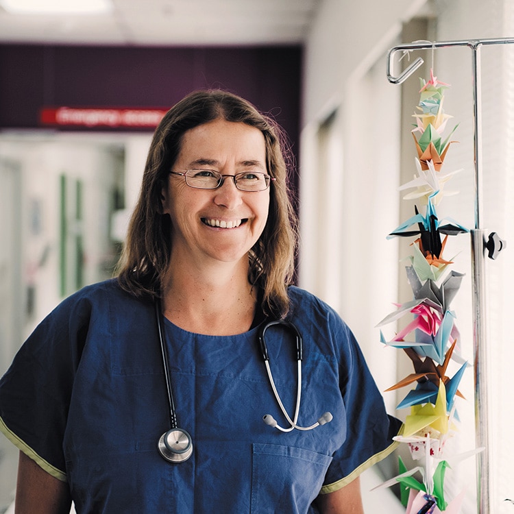 Catherine Crock stands in blue hospital scrubs smiling off-camera, next to an IV stand with a string of coloured paper cranes.
