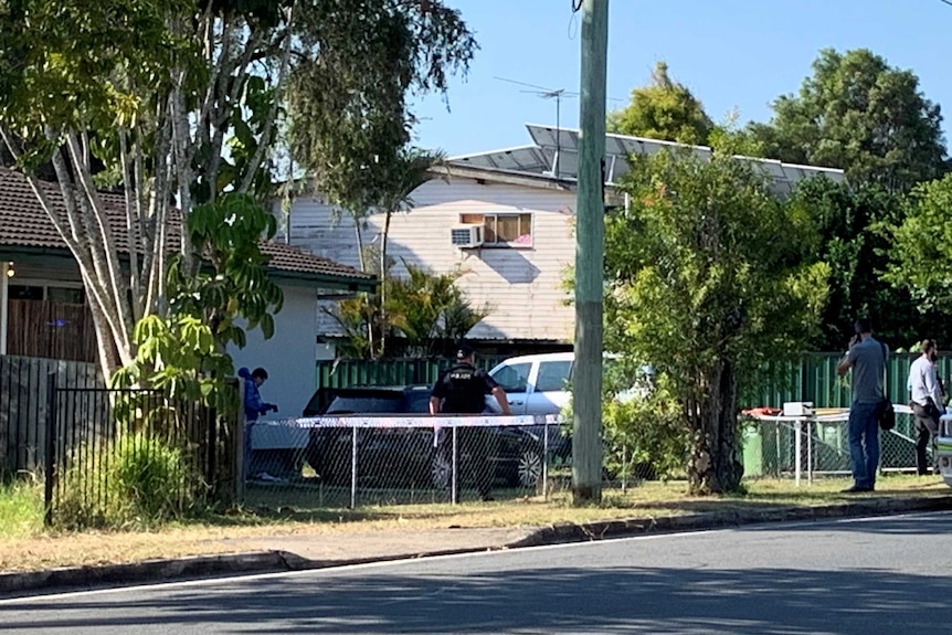 A policeman is seen behind a fence, with a man in a blue hoodie to his left with a camera. Two further men are to the right.