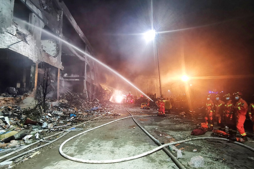 A group of people in orange overalls and face masks hose down a gutted building at night time.