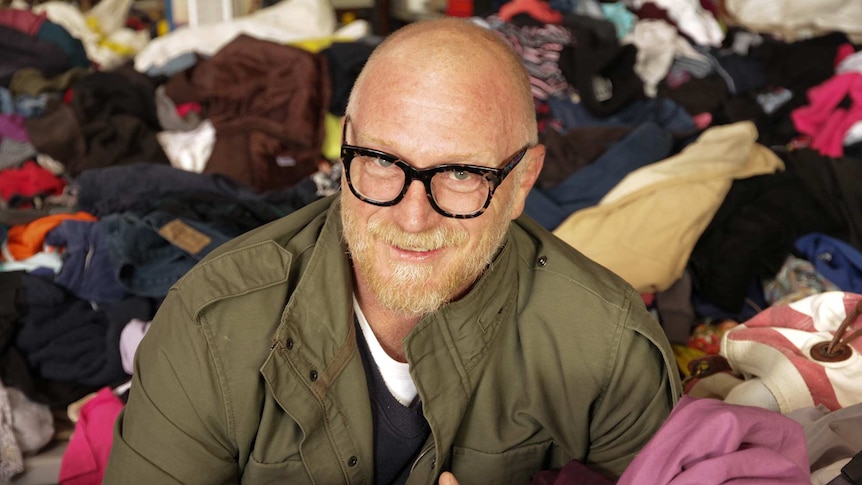 A man with black spectacles pictured surrounded by mountains of second hand clothes.