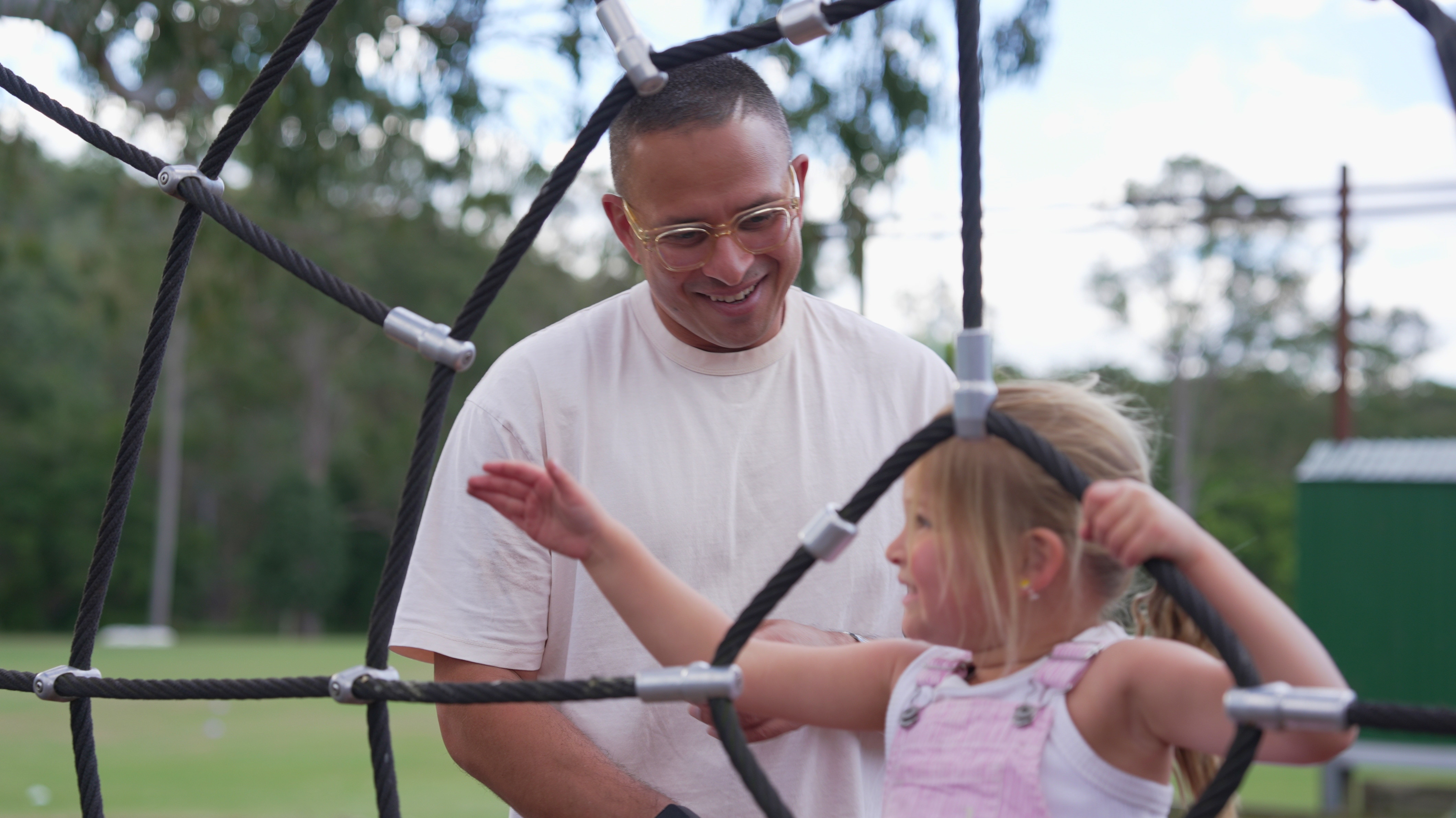 Usman Khawaja, wearing glasses, looks down at Ayla and smiles as she holds onto play equipment.