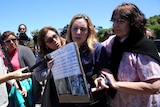 A woman speaks to media holding up a sign with her husband's name on it.