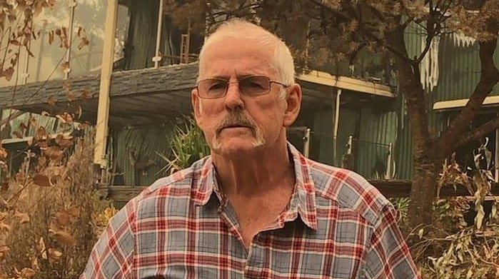 An older man stands in front of a house that has been destroyed by fire.