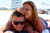 Man in sunglasses lies on beach, woman in blue bikini rests of top of him with one hand covering face